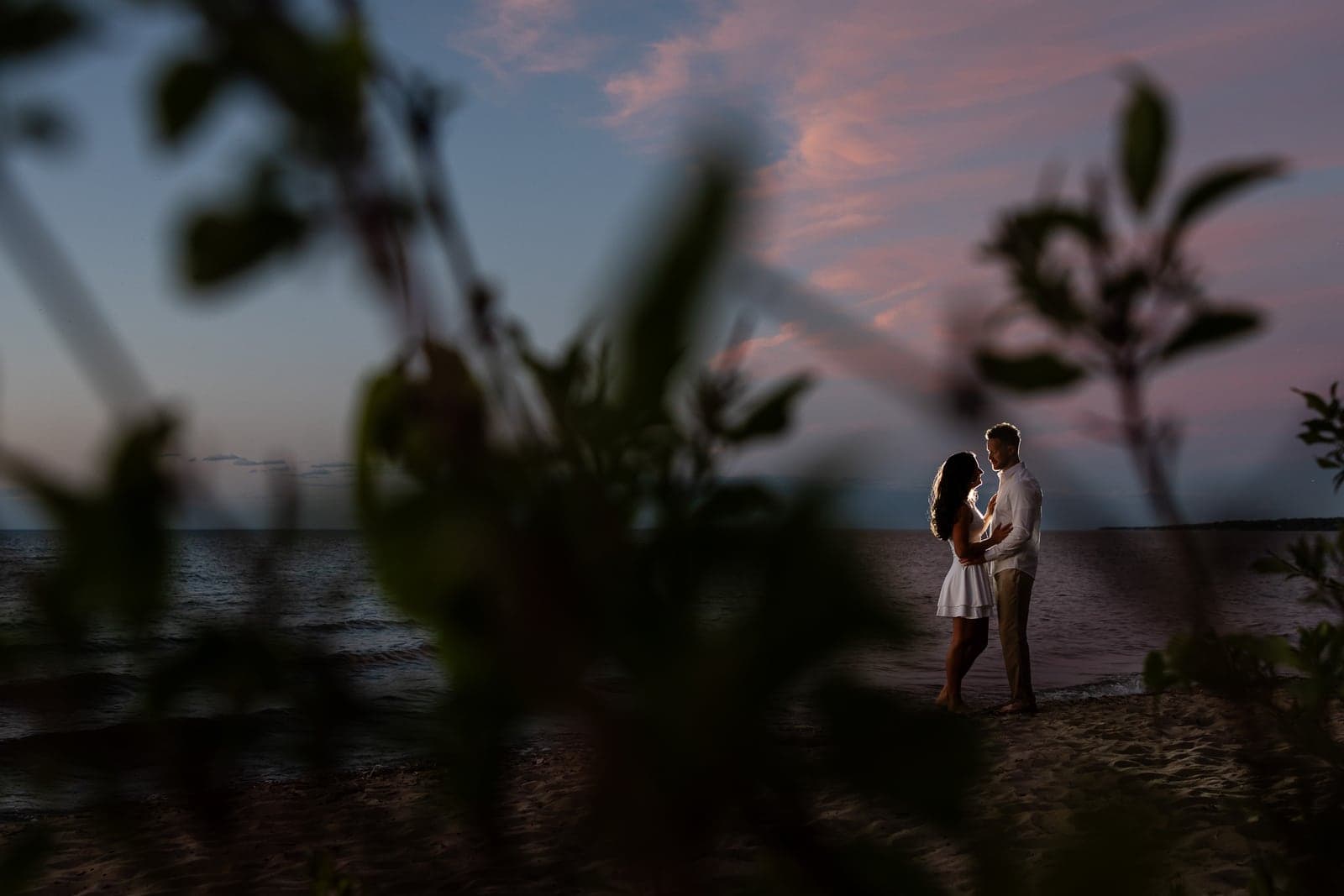 Danielle and Sheldon facing each other on the beach at dusk, framed by blurred leaves